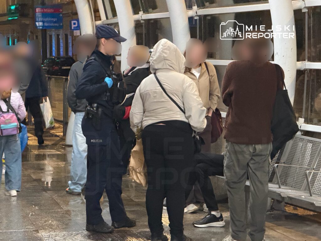 Policjant w mundurze rozmawia z grupą osób na przystanku, w tle widać deszczową nawierzchnię i znaki drogowe.