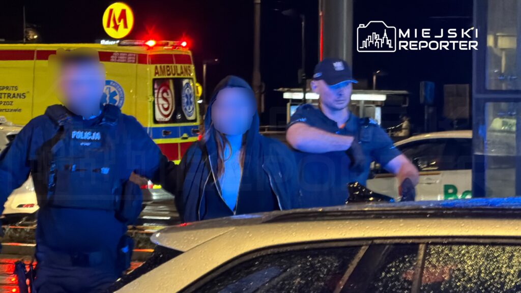 Policjanci zatrzymują osobę w ciemnym ubraniu w pobliżu karetki pogotowia na oświetlonej ulicy.