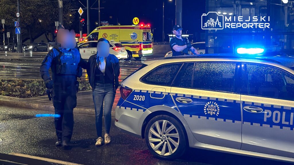 Policjant prowadzi kobietę w dżinsach i kurtce do radiowozu w deszczowy wieczór na ulicy.