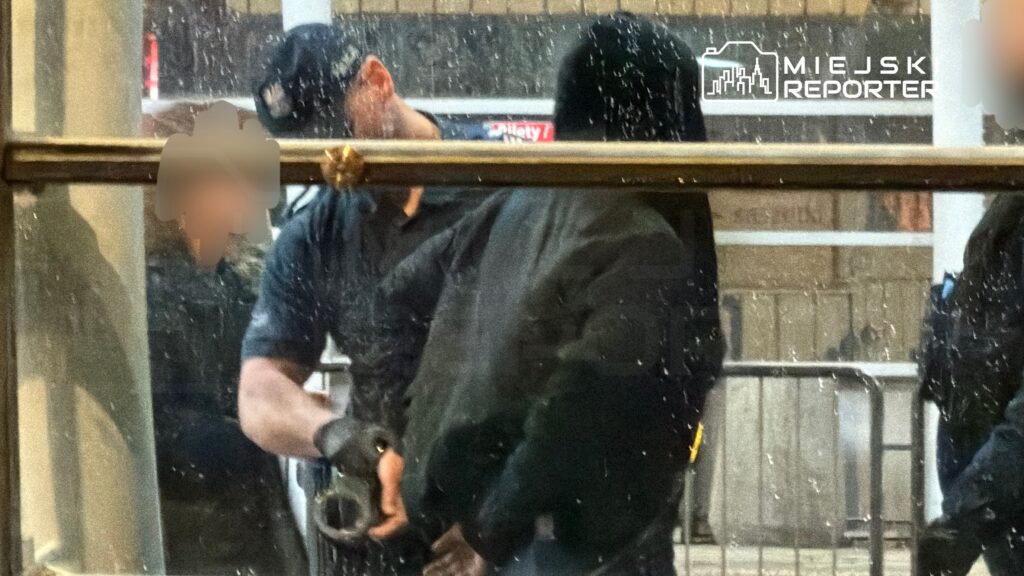 Funkcjonariusz policji aresztuje osobę w ciemnej odzieży w deszczowym otoczeniu.