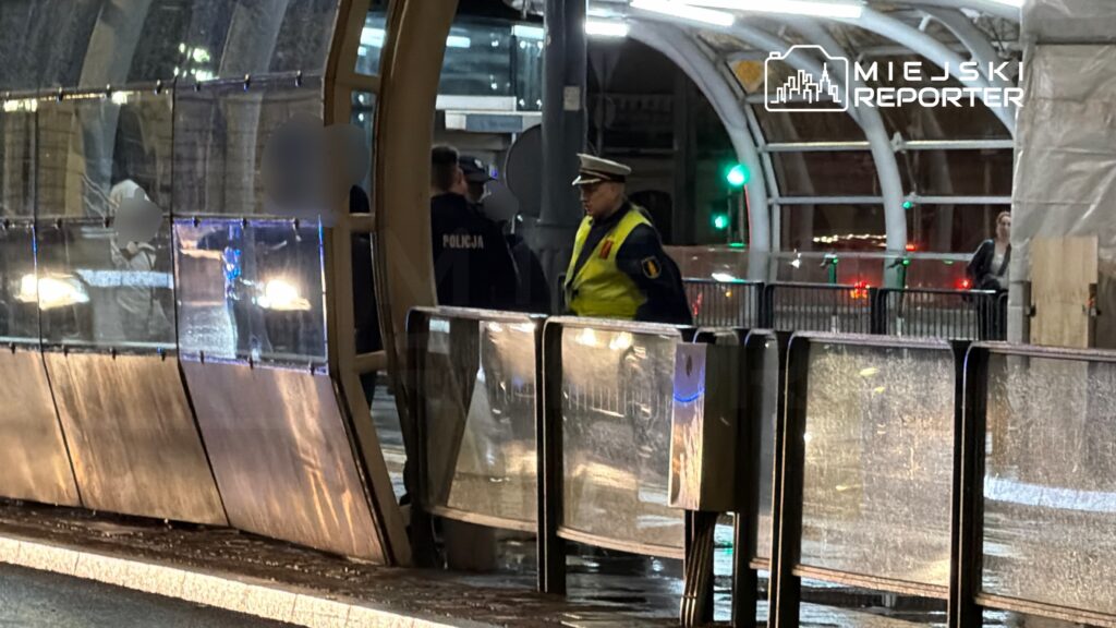 Policjant w żółtej kamizelce kontroluje ruch na przystanku tramwajowym w nocy, w tle widać pojazdy.