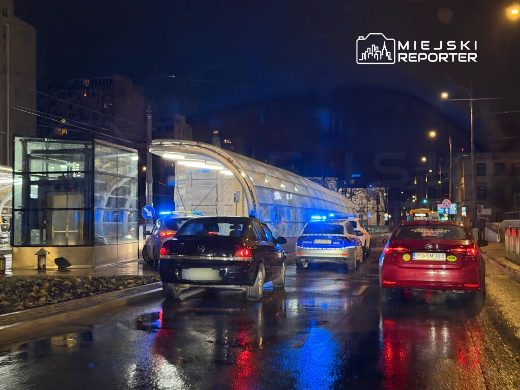 Policyjny radiowóz z włączonymi sygnałami świetlnymi zatrzymany przy przystanku tramwajowym w nocy. W tle widać oświetlone budynki.