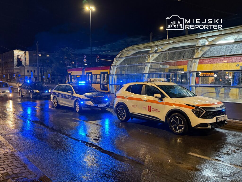 Policyjne samochody z niebieskimi światłami stoją na mokrej ulicy, obok przystanku tramwajowego w nocy.