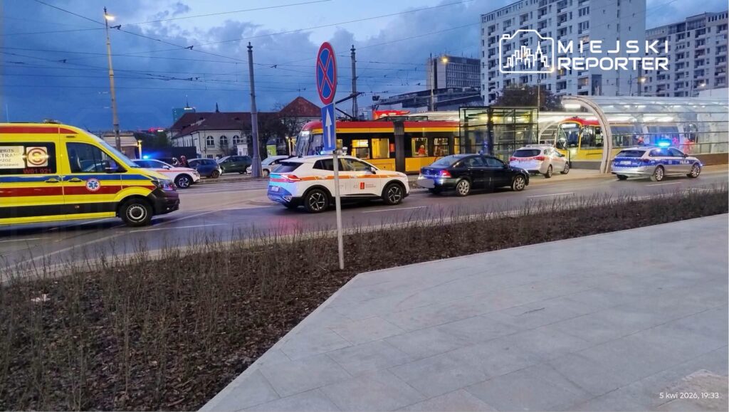 Karetka pogotowia oraz radiowozy policyjne zatrzymane na ulicy obok przystanku tramwajowego.