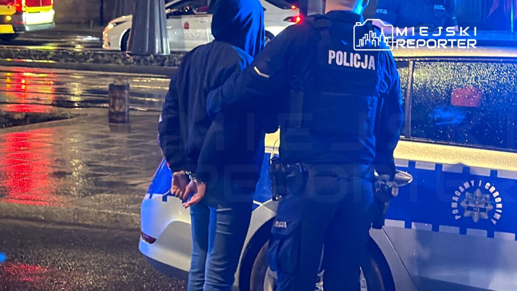 Mężczyzna w ciemnym ubraniu jest zatrzymywany przez policjanta w mundurze w deszczowej noc na ulicy.