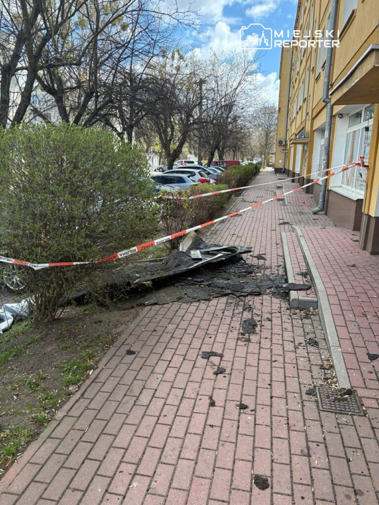 Zniszczony fragment chodnika z rozdartą nawierzchnią, ogrodzony taśmą ostrzegawczą, w otoczeniu drzew i parkujących samochodów.
