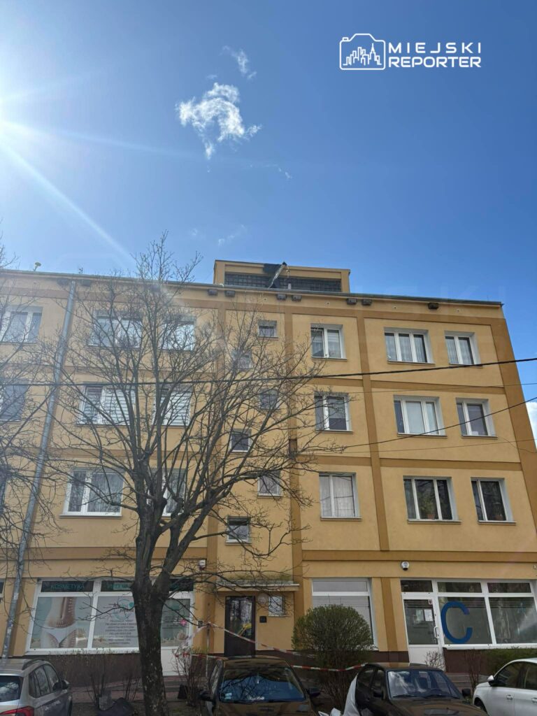 Czteropiętrowy budynek mieszkalny w żółtej elewacji z drzewem na pierwszym planie oraz niebieskim niebem w tle.