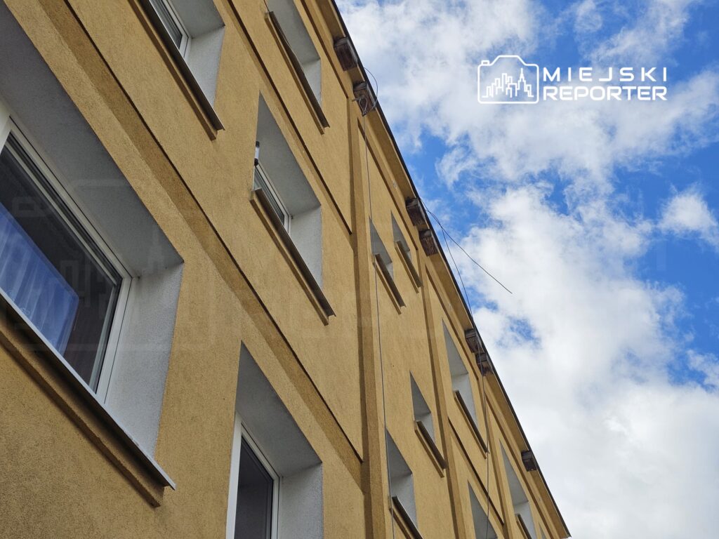 Żółty budynek mieszkalny z oknami i fragmentem nieba w tle, z białymi chmurami.