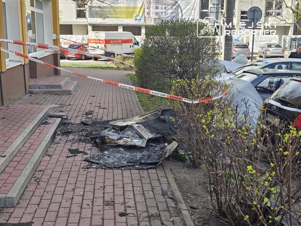 Zniszczony kawałek chodnika z czarnym gruzem obok budynku, otoczony taśmą policyjną, w tle zaparkowane samochody.