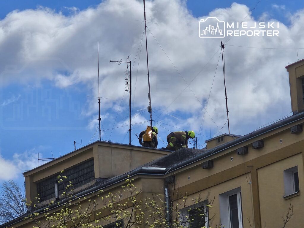 Dwaj pracownicy w kaskach montują anteny na dachu budynku w słoneczny dzień, z chmurami na tle.