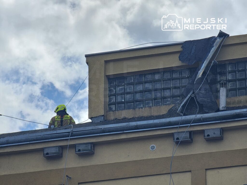 Strażak w żółtej kasku przegląda uszkodzenie dachu budynku, na którym widoczne są fragmenty zerwanego pokrycia.