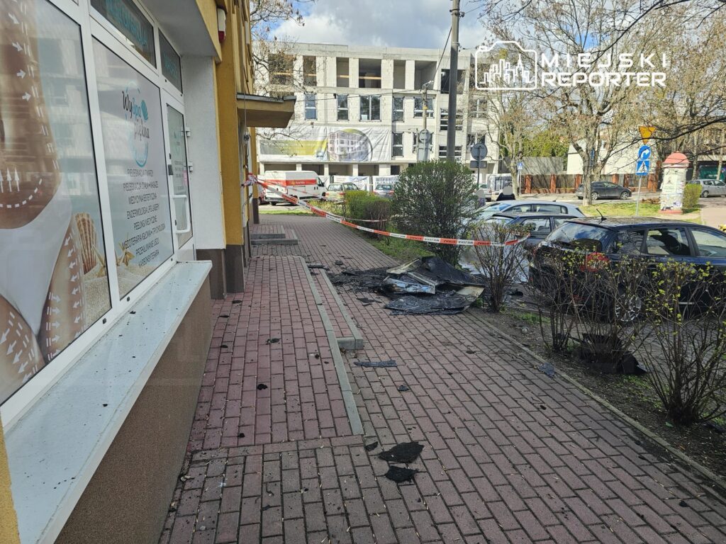 Zniszczona nawierzchnia chodnika z resztkami czarnego materiału, otoczona taśmą zabezpieczającą, obok nowego budynku.