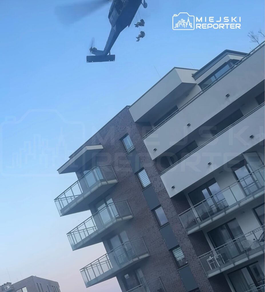 Helikopter wojskowy z żołnierzami na linach przelatuje nad nowoczesnym budynkiem mieszkalnym.
