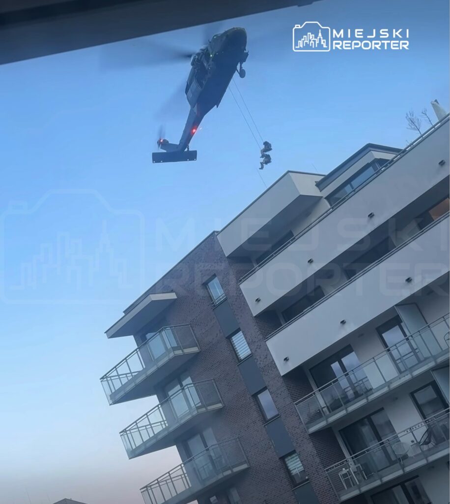 Śmigłowiec policyjny unosi się nad nowoczesnym budynkiem mieszkalnym, a dwóch ratowników zjeżdża na linie.
