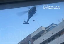 Helikopter z opadającym ratownikiem przelatuje nad nowoczesnym budynkiem mieszkalnym.