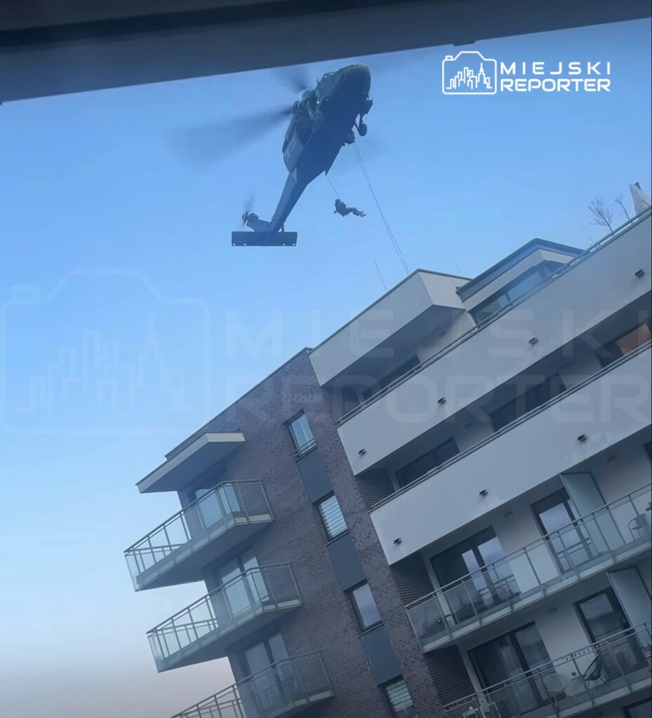 Helikopter z opadającym ratownikiem przelatuje nad nowoczesnym budynkiem mieszkalnym.