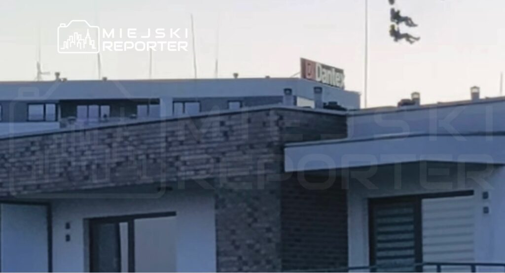 Dwie postacie w sprzęcie spadochronowym zjeżdżają na linach z dachu budynku. W tle widoczny napis "Dantex".