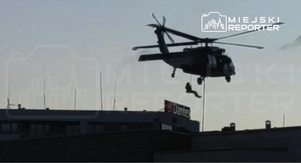 Helikopter ratunkowy unosi się nad dachem budynku, z którego zwisa osoba na linie.