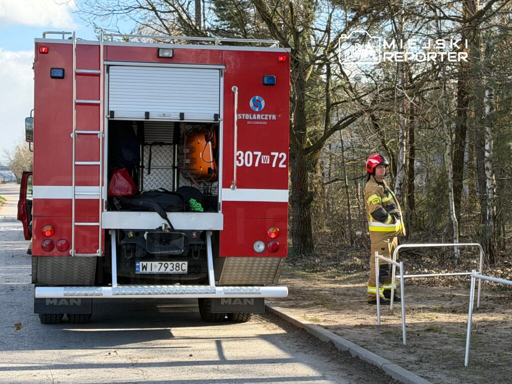 Strażak w hełmie stoi obok wozu strażackiego na leśnej drodze, przygotowując się do akcji.