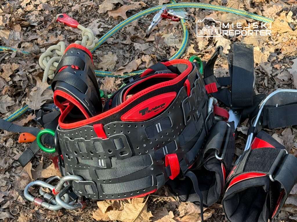 Czarny pas wspinaczkowy z czerwonymi akcentami leży na liściach, obok lin i karabinków.