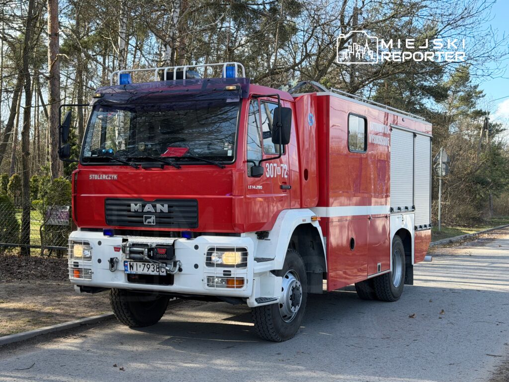 Czerwony wóz strażacki marki MAN zaparkowany na drodze w otoczeniu lasu, z widocznymi reflektorami i sygnalizacją świetlną.