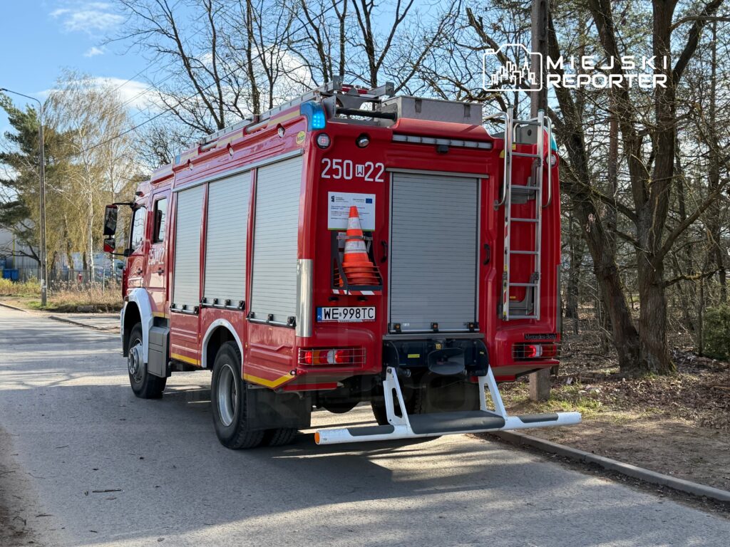 Czerwony wóz strażacki zaparkowany na poboczu drogi, obok drzew i budynków. Na dachu widać niebieskie światło.