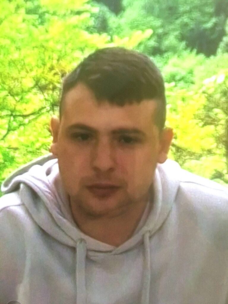 Mężczyzna w szarej bluzie siedzi w otoczeniu zieleni, na tle drzew i krzewów.