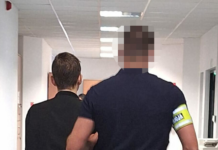 Mężczyzna w czarnym ubraniu prowadzony przez mężczyznę w niebieskiej koszulce z żółtą opaską w biurowym korytarzu.