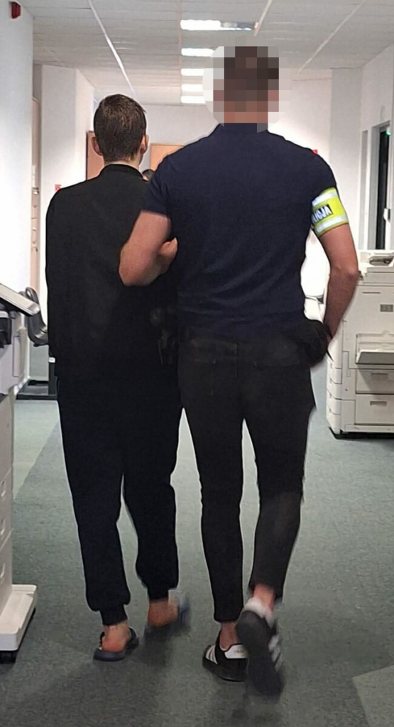 Mężczyzna w czarnym ubraniu prowadzony przez mężczyznę w niebieskiej koszulce z żółtą opaską w biurowym korytarzu.