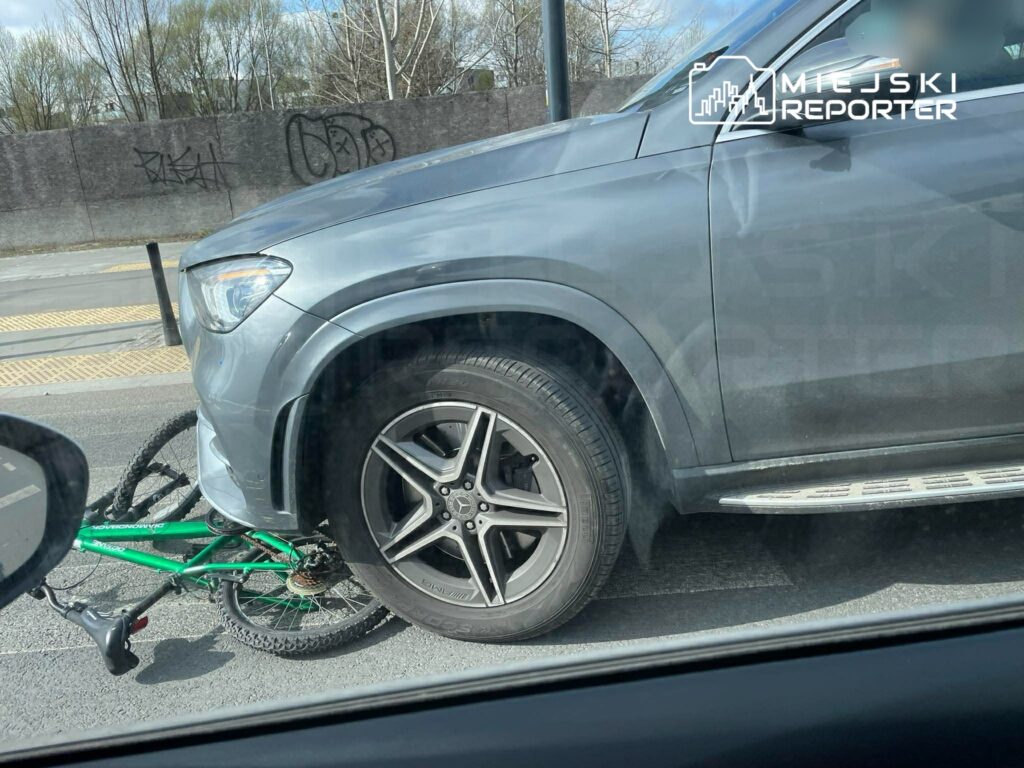 Zielony rower leży pod przednim kołem szarego SUV-a na ruchliwej ulicy, w tle widoczny mur z graffiti.