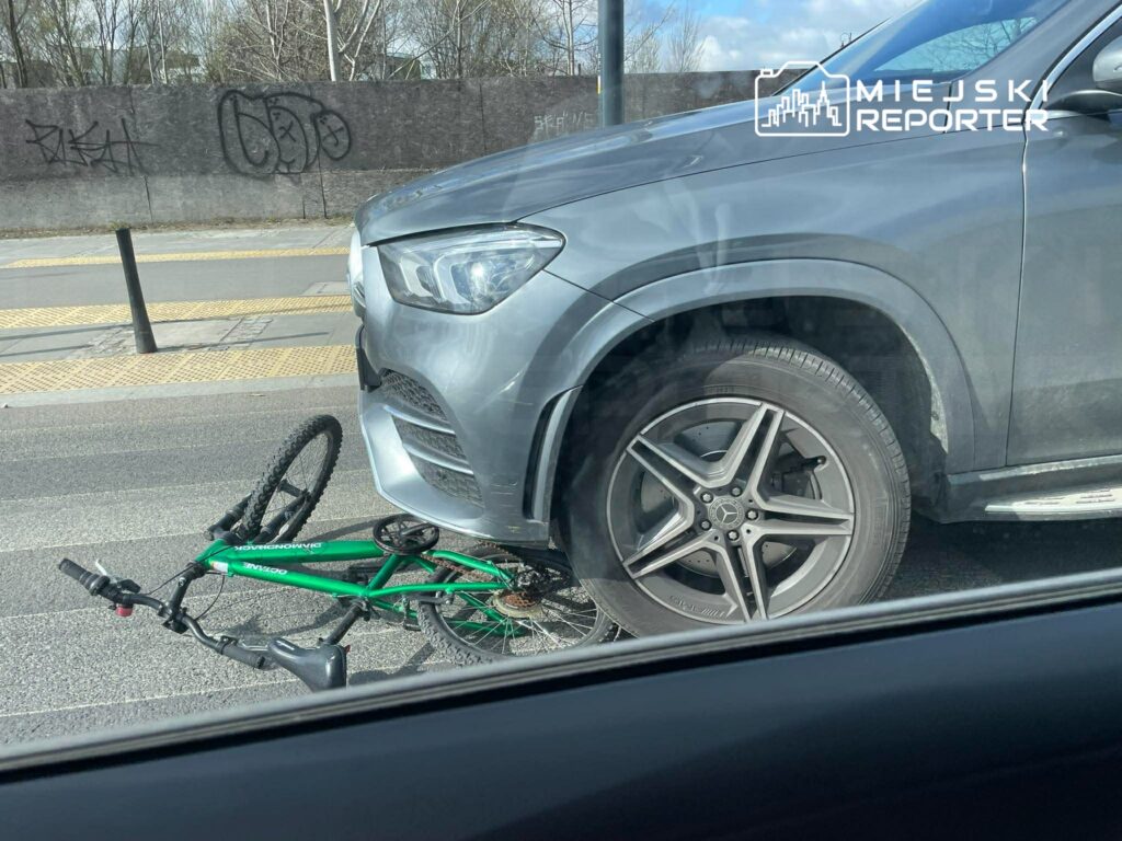 Zielony rower leży pod przednim kołem szarego SUV-a na miejskiej ulicy.