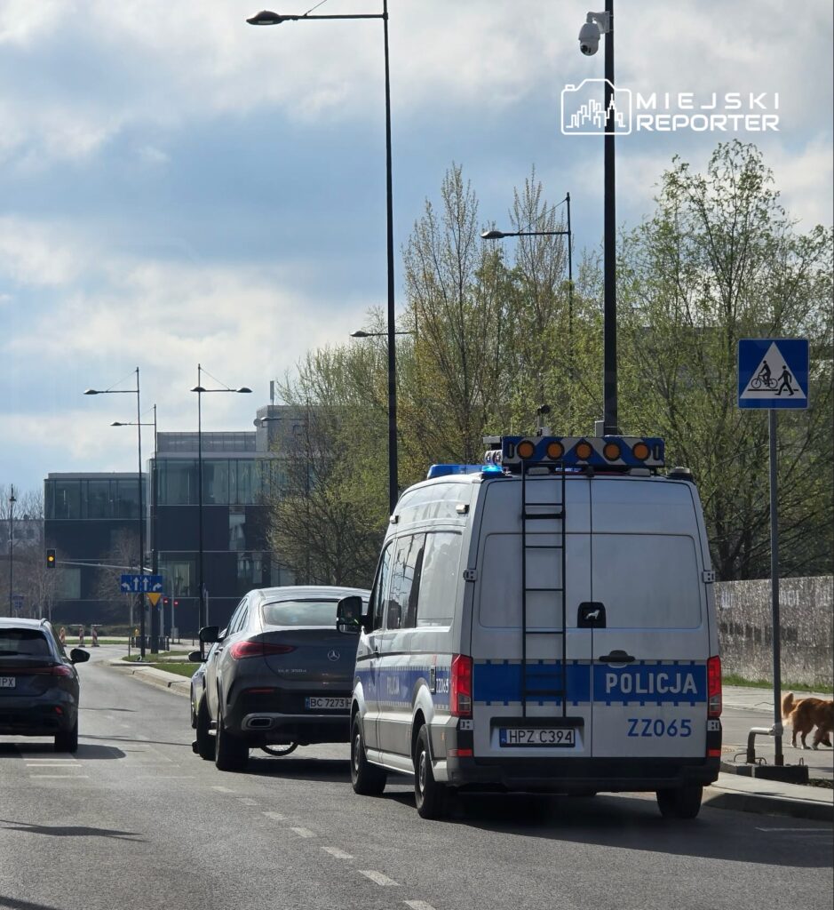 Policyjny furgon zatrzymany na ulicy, obok osobowy samochód marki Mercedes, w tle nowoczesny budynek biurowy.