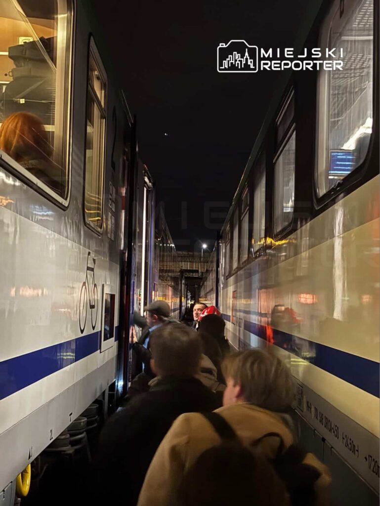 Grupa pasażerów wchodzi do pociągu na stacji kolejowej w nocy, między dwoma wagonami.