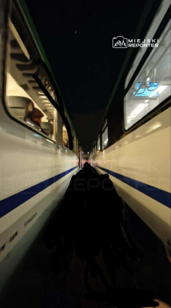 Dwa pociągi stoją blisko siebie na stacji, otoczone nocnym mrokiem. Widać wnętrza wagonów przez okna.