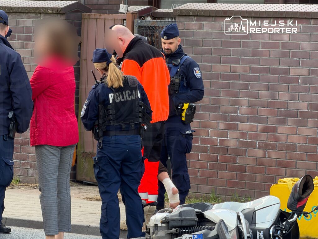 Grupa policjantów rozmawia z kobietą w różowej kurtce, obok stoi funkcjonariusz w pomarańczowej odzieży roboczej.