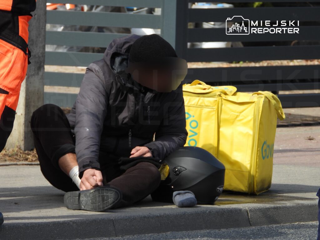 Mężczyzna w czarnej kurtce siedzi na chodniku, dotykając stopy, obok żółte torby dostawcze.