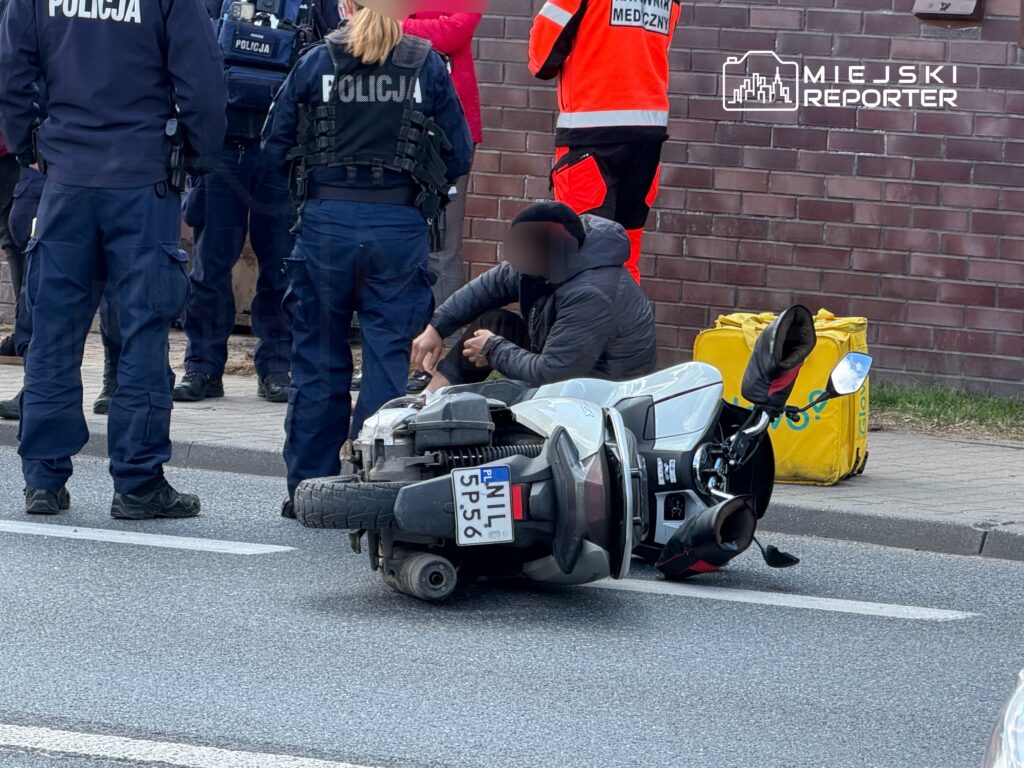 Mężczyzna przywracający motocykl do pionu na jezdni, w otoczeniu policjantów i ratowników medycznych.