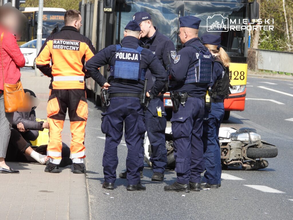 Ratownik medyczny i policjanci udzielają pomocy poszkodowanemu na ulicy obok przewróconego motocykla.