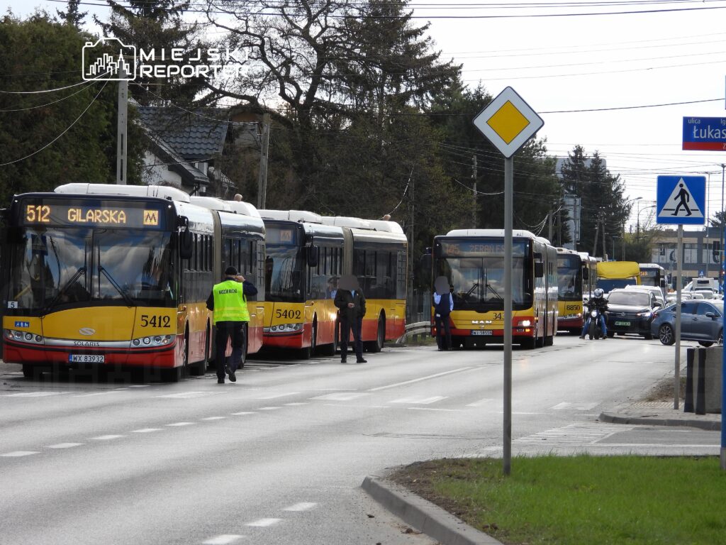 Trzy autobusy komunikacji miejskiej zatrzymane na ulicy, przy których pracownicy w kamizelkach odblaskowych pomagają pasażerom.