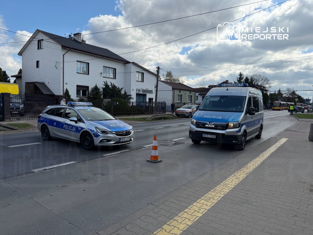 Policyjny radiowóz i furgonetka wstrzymują ruch na ulicy, obok białego budynku mieszkalnego.