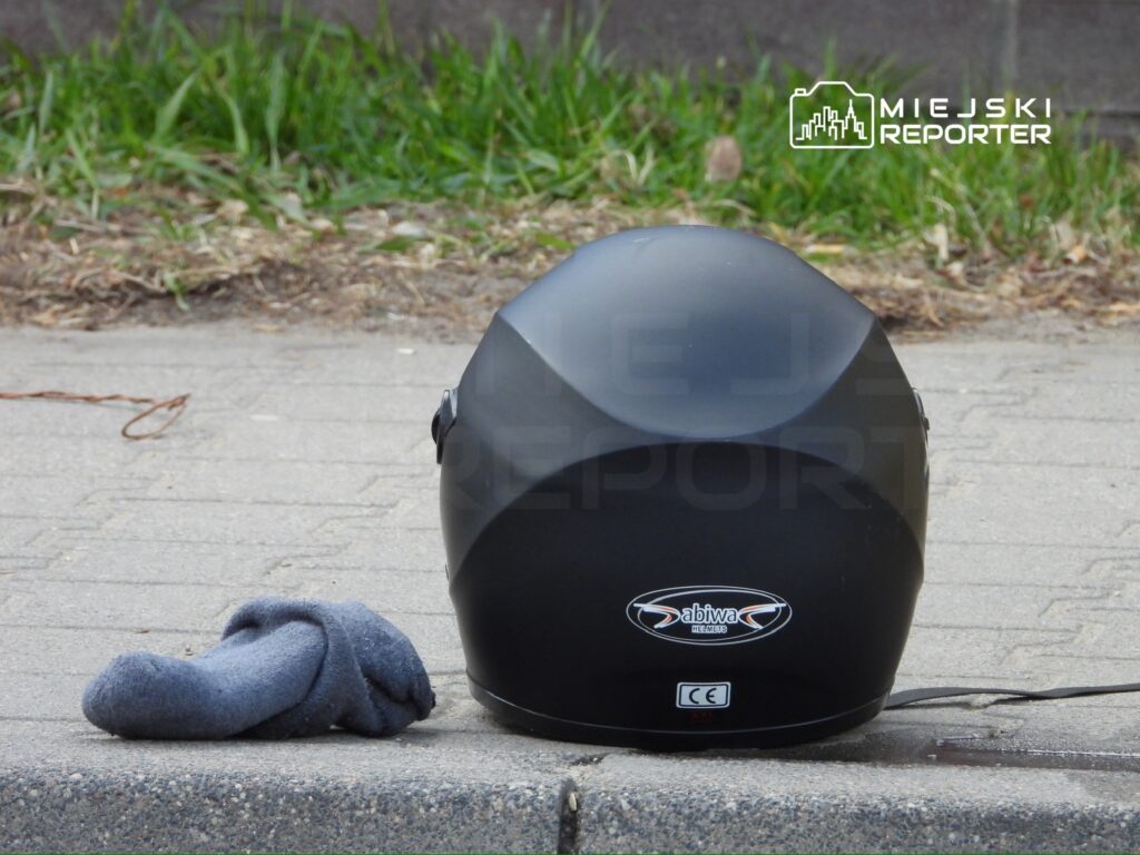Czarny kask motocyklowy leży na chodniku obok zmiętej niebieskiej skarpetki, otoczony trawą.