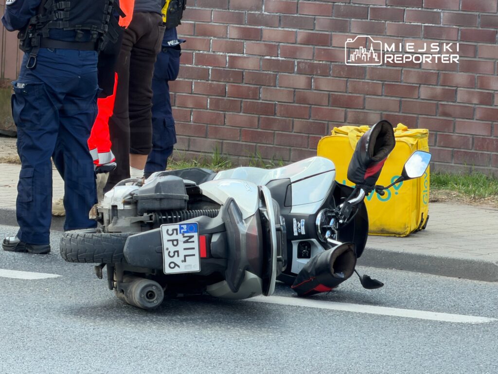 Na drodze leży przewrócony skuter, obok grupa osób w mundurach policyjnych oraz ratowników medycznych.