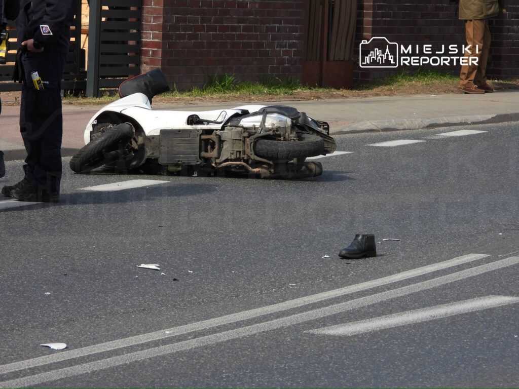 Policjant bada przewrócony skuter leżący na jezdni obok zgubionego buta. W tle widoczna czerwona ceglana ściana.