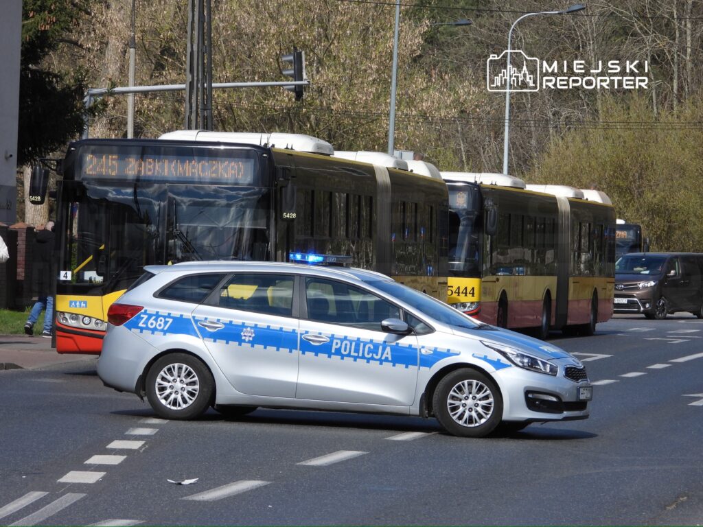 Policyjny radiowóz blokuje ruch na ulicy, obok zatrzymane autobusy miejskie z wyświetlaczami tras.