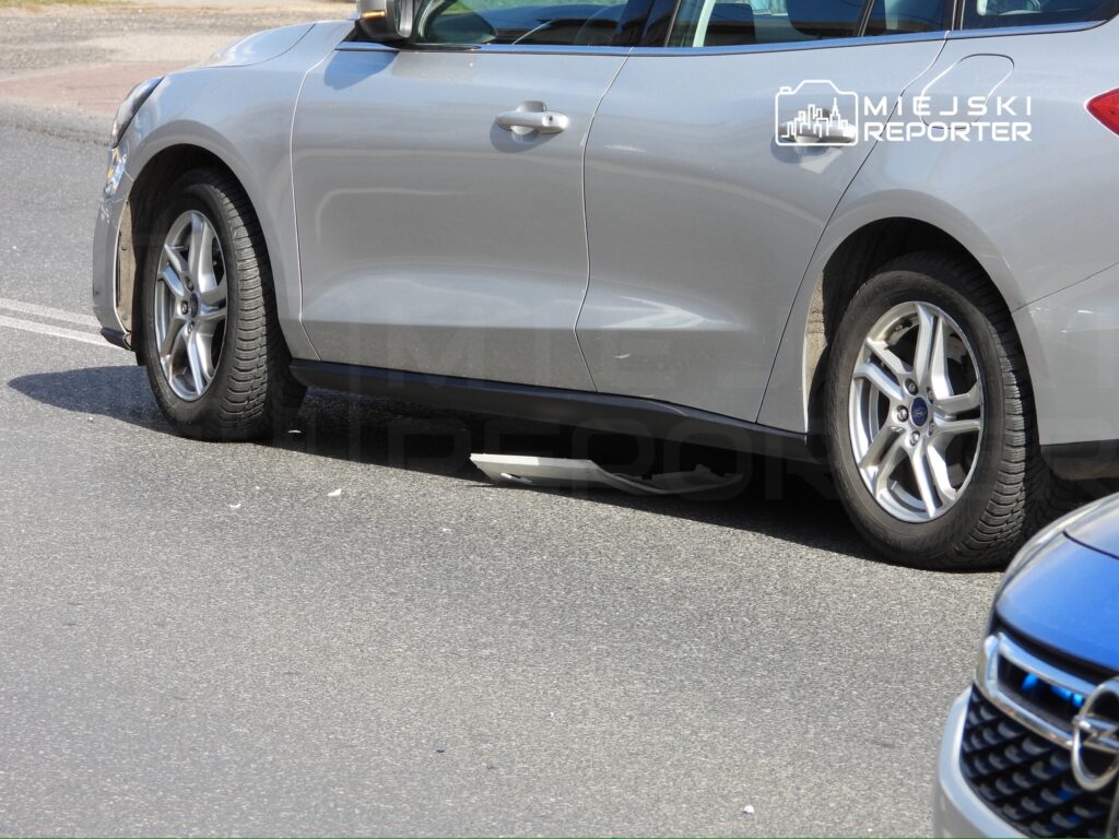 Srebrny samochód osobowy z uszkodzoną osłoną pod silnikiem zaparkowany na drodze.