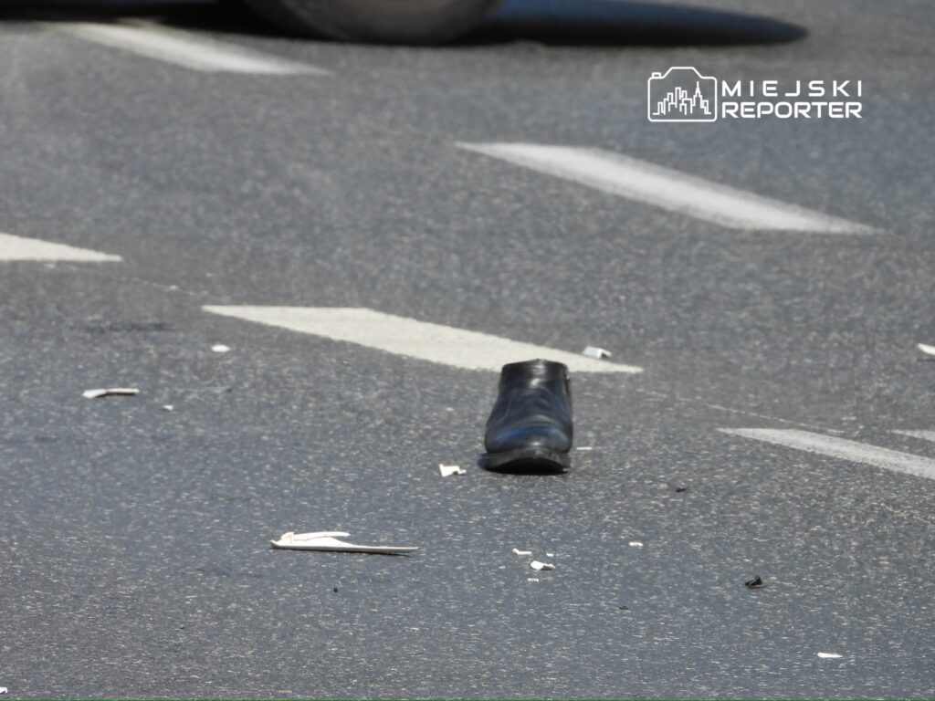 Na jezdni leży czarny but, obok widoczne fragmenty papierów i zarysowane pasy drogowe.