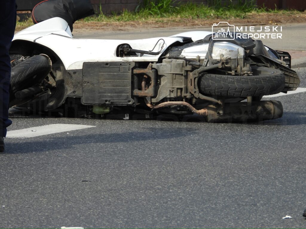 Motocykl leży na boku na drodze, obok stoi policjant w mundurze, badając sytuację.