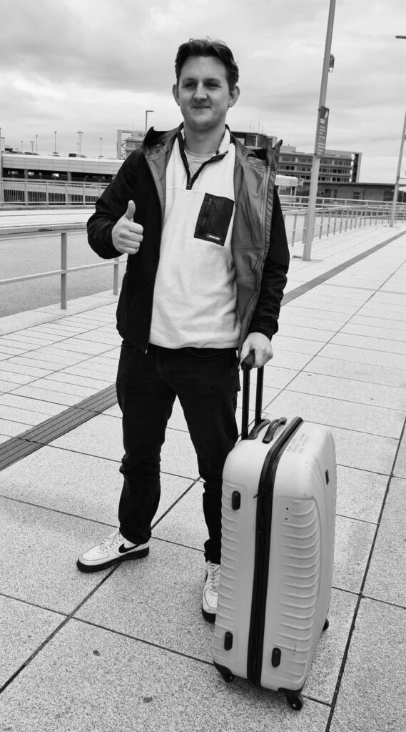 Mężczyzna z walizką stoi na chodniku, uśmiechając się i pokazując kciuk w górę.