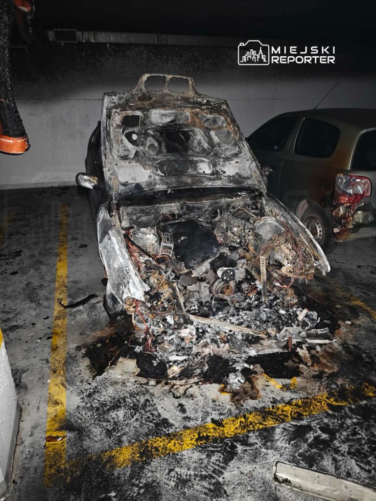 Spalony wrak samochodu osobowego w podziemnym parkingu, otoczony zniszczoną posadzką i śladami po pożarze.