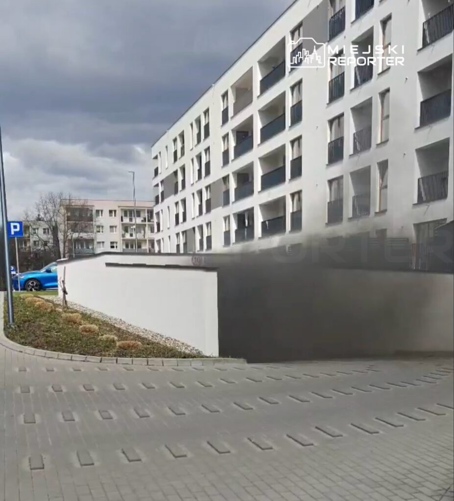 Nowoczesny budynek mieszkalny z balkonami, w tle niebieski samochód zaparkowany w strefie parkingowej.